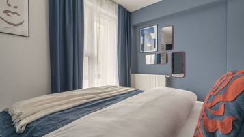 Un dormitorio con cama, ventana con cortinas, radiador y un espejo y obras de arte en la pared.
