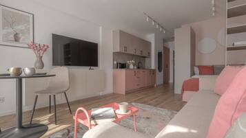 A modern living room with a sofa, a coffee table, and a TV. There is a kitchen area with pink cabinets and a dining table with chairs.