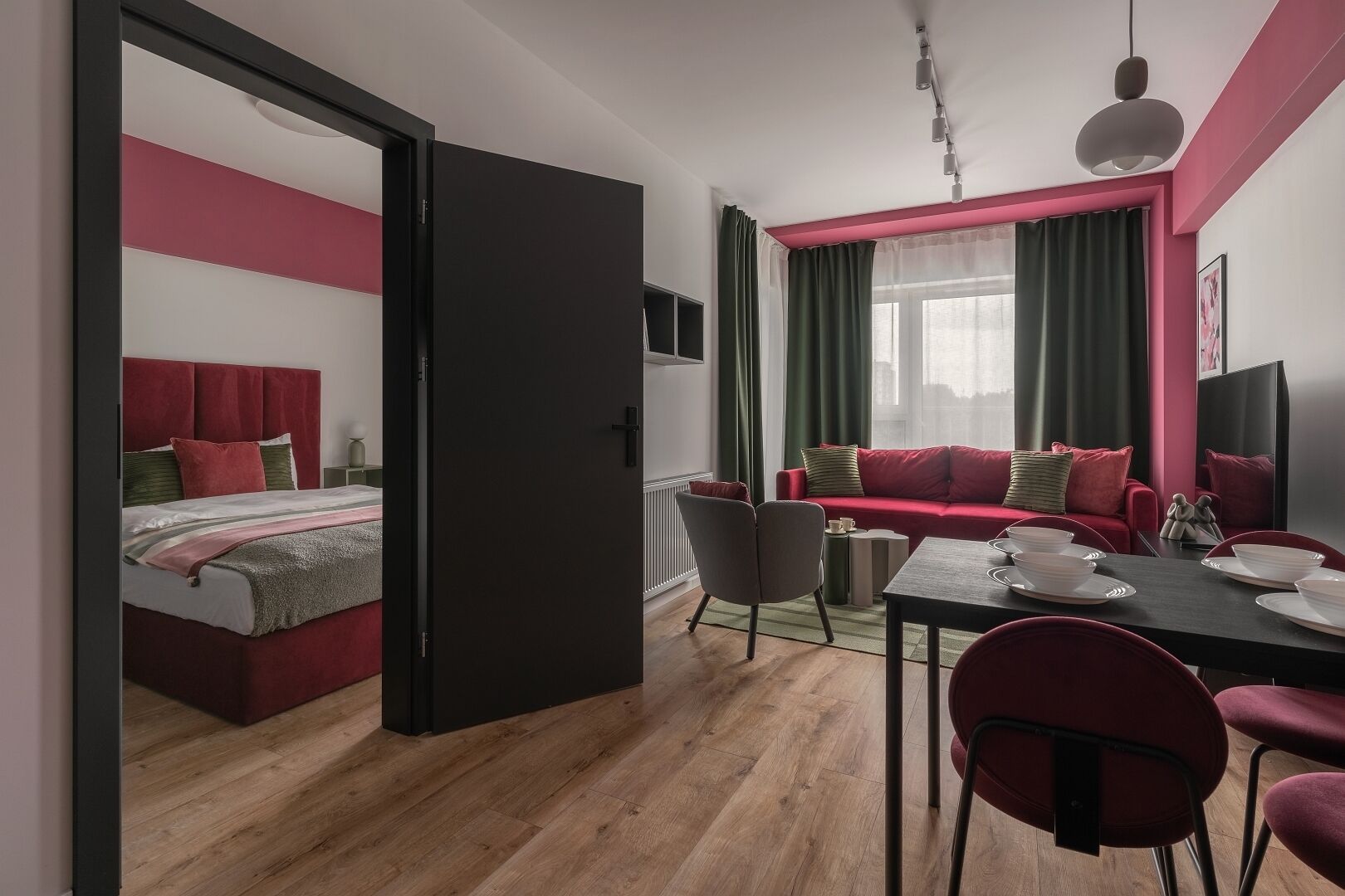 A modern, well-lit room with a bed, a red sofa, a dining table set for two, and a large window with curtains.