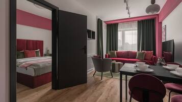 A modern, well-lit room with a bed, a red sofa, a dining table set for two, and a large window with curtains.