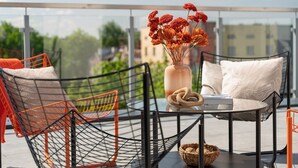 A modern outdoor seating area with a round glass table, orange mesh chairs, and a vase with orange flowers. - Apartio Rooms Zabrze (Zabrze)