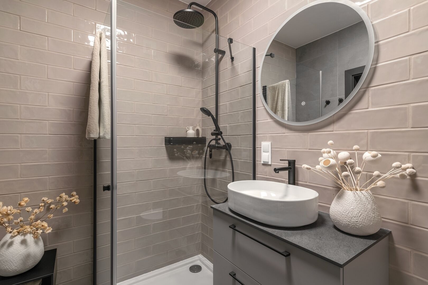 Une salle de bain moderne avec une douche à parois de verre, un miroir rond, un lavabo blanc et un vase contenant des fleurs séchées.