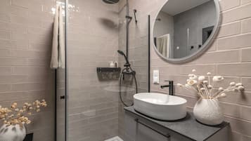 Une salle de bain moderne avec une douche à parois de verre, un miroir rond, un lavabo blanc et un vase contenant des fleurs séchées.