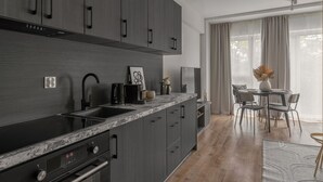 A modern kitchen with dark cabinetry, a marble countertop, and a built-in oven. There is a dining area with a table and chairs, and a large window with curtains. - Apartio Rooms Zabrze (Zabrze)