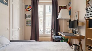 Habitación de hotel con cama, escritorio, computadora, silla, ventana con cortinas y radiador.