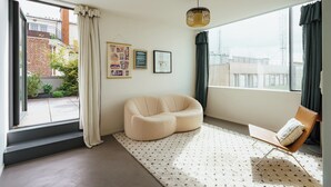 A modern living room with a curved sofa, a small wooden chair, and a patterned rug. There are two framed pictures on the wall and a large window with curtains.