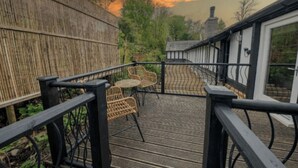 Eine Holzterrasse mit Korbstühlen und einem Tisch, mit Blick auf einen Garten und ein Gebäude mit Schornstein.