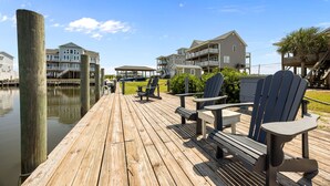 A wooden deck with Adirondack chairs and a covered seating area, overlooking a body of water with buildings in the background. - Waterfront Paradise in N. Topsail: Ocean/Canal Views, Fishing, Steps from Beach (North Topsail Beach)