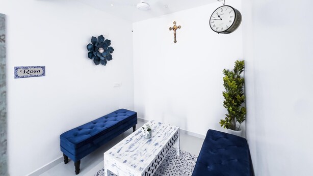 Ein kleiner Raum mit weißen Wänden, einer blauen Bank, einem weißen Tisch mit blauem Muster, einer Uhr, einem Kreuz und einer Pflanze.