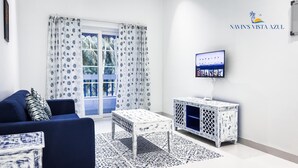 Un salon avec un canapé bleu, des rideaux à motifs, un meuble blanc fixé au mur sur lequel est posée une télévision, et un tapis à motifs.