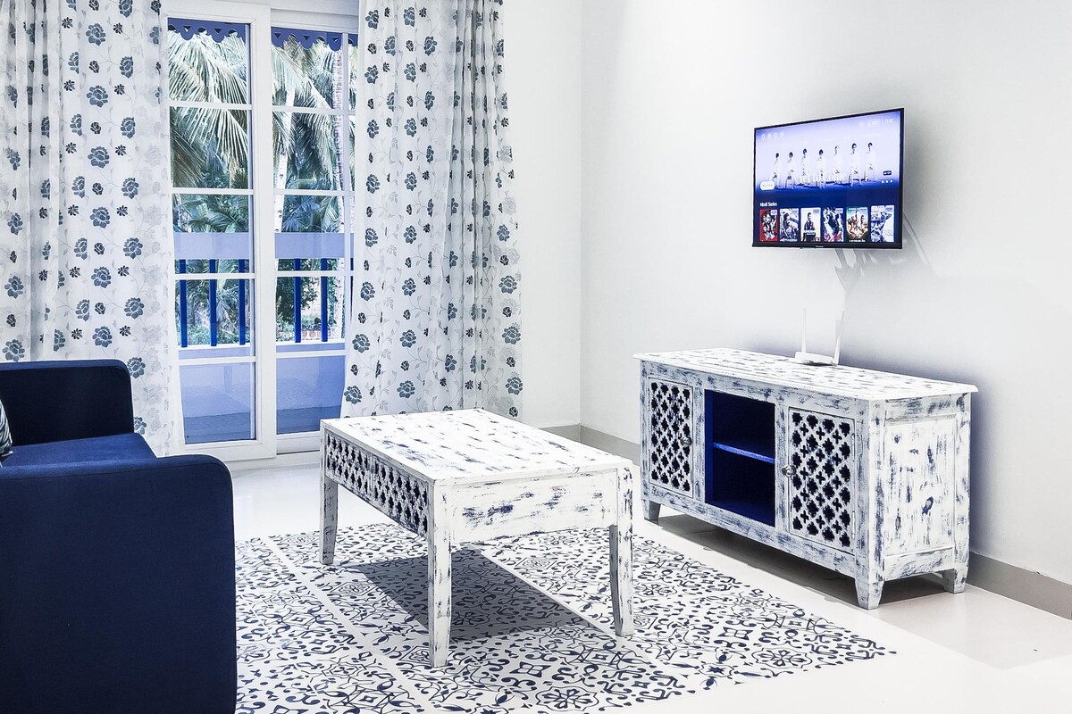 Ein Wohnzimmer mit einem blauen Sofa, gemusterten Vorhängen, einem weißen Schrank und einem Fernseher an der Wand sowie einem gemusterten Teppich.