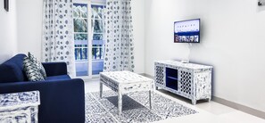 Ein Wohnzimmer mit einem blauen Sofa, gemusterten Vorhängen, einem weißen Schrank und einem Fernseher an der Wand sowie einem gemusterten Teppich.