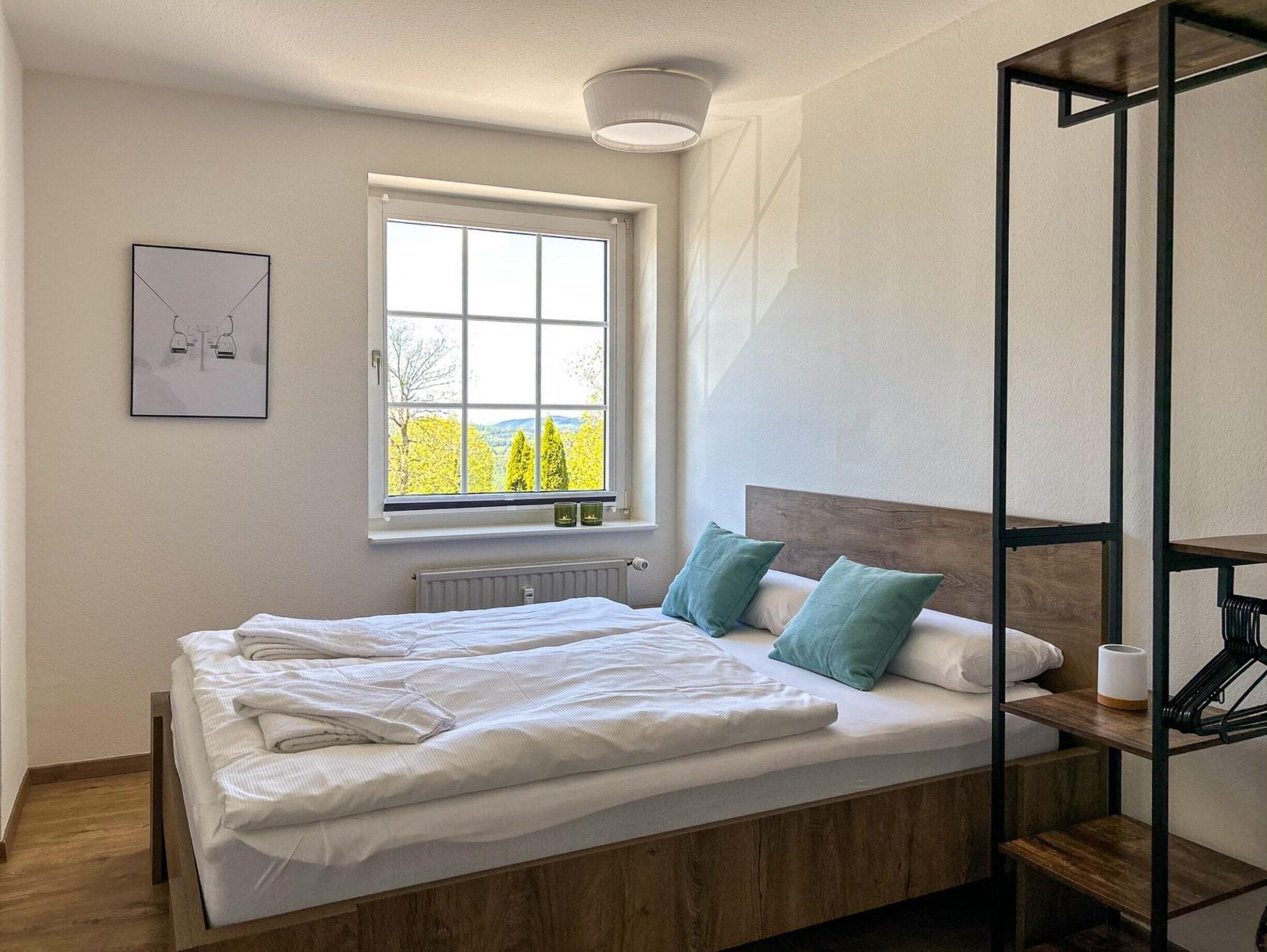 Un lit bien fait, avec du linge de lit blanc, une vue sur les arbres par la fenêtre et une petite étagère avec une lampe et un rouleau de papier toilette.