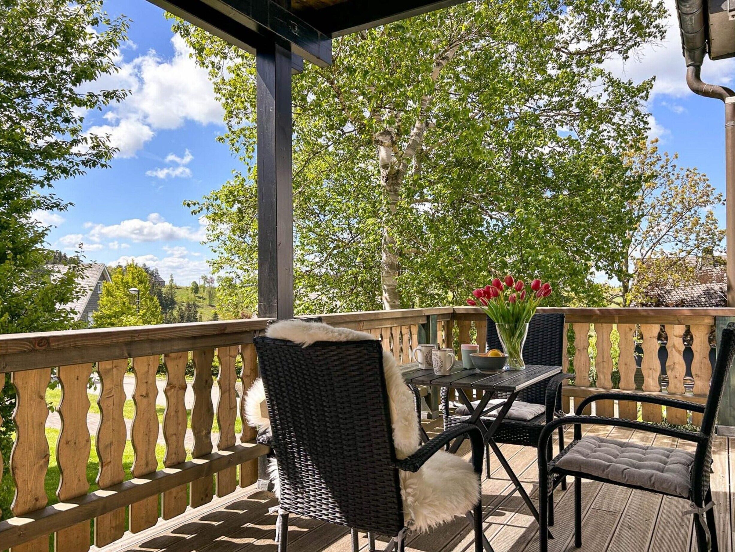 A covered outdoor seating area with wicker chairs and a table, surrounded by a wooden railing and trees.