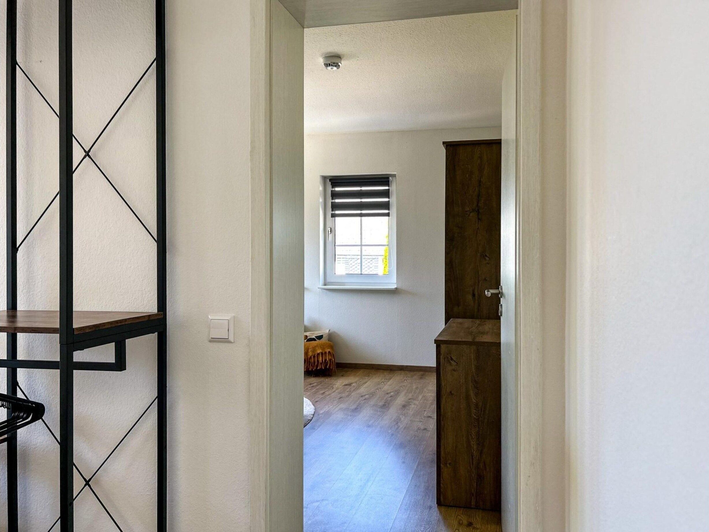 A narrow hallway leading to a room with a window, a wooden door, and a light switch on the wall.