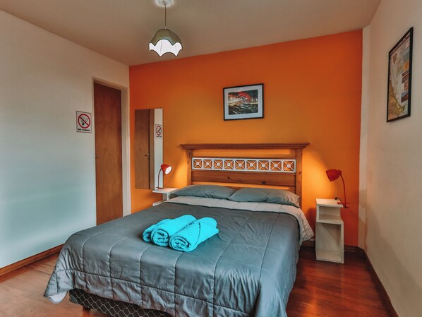Een slaapkamer met een bed, een nachtkastje, een lamp, een aan de muur hangend schilderij, en een deur met een 'Niet roken'-bordje.