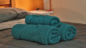 A neatly made bed with two rolled-up teal towels and a red desk lamp in the background.