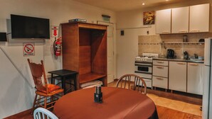 A compact kitchen with white cabinets, a stove, and a refrigerator. There's a dining table with chairs and a small black table. A fire extinguisher and a "No Smoking" sign are visible on the wall.