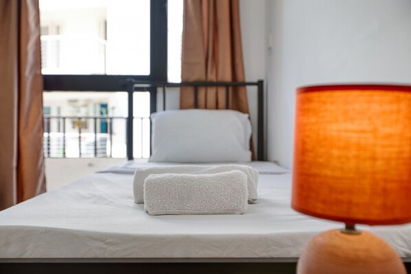 A bed with white linens and two folded towels, a window with curtains, and a lamp with a textured shade.