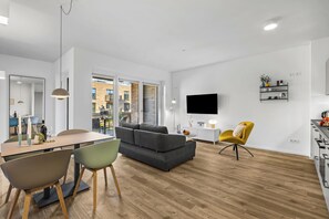 A modern living room with a grey sofa, a dining area with a table and chairs, a yellow armchair, and a TV mounted on the wall. - Modern apartment with terrace, partial sea view and private parking in Travemünd (Lübeck)