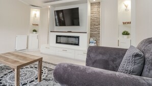 A modern living room with a flat-screen TV mounted above a fireplace, white cabinetry below, and a grey sofa. - The Cabin (Malton)