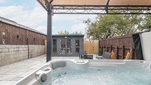 A hot tub in a backyard with a wooden fence, a covered patio, and a small building with glass doors. - The Cabin (Malton)