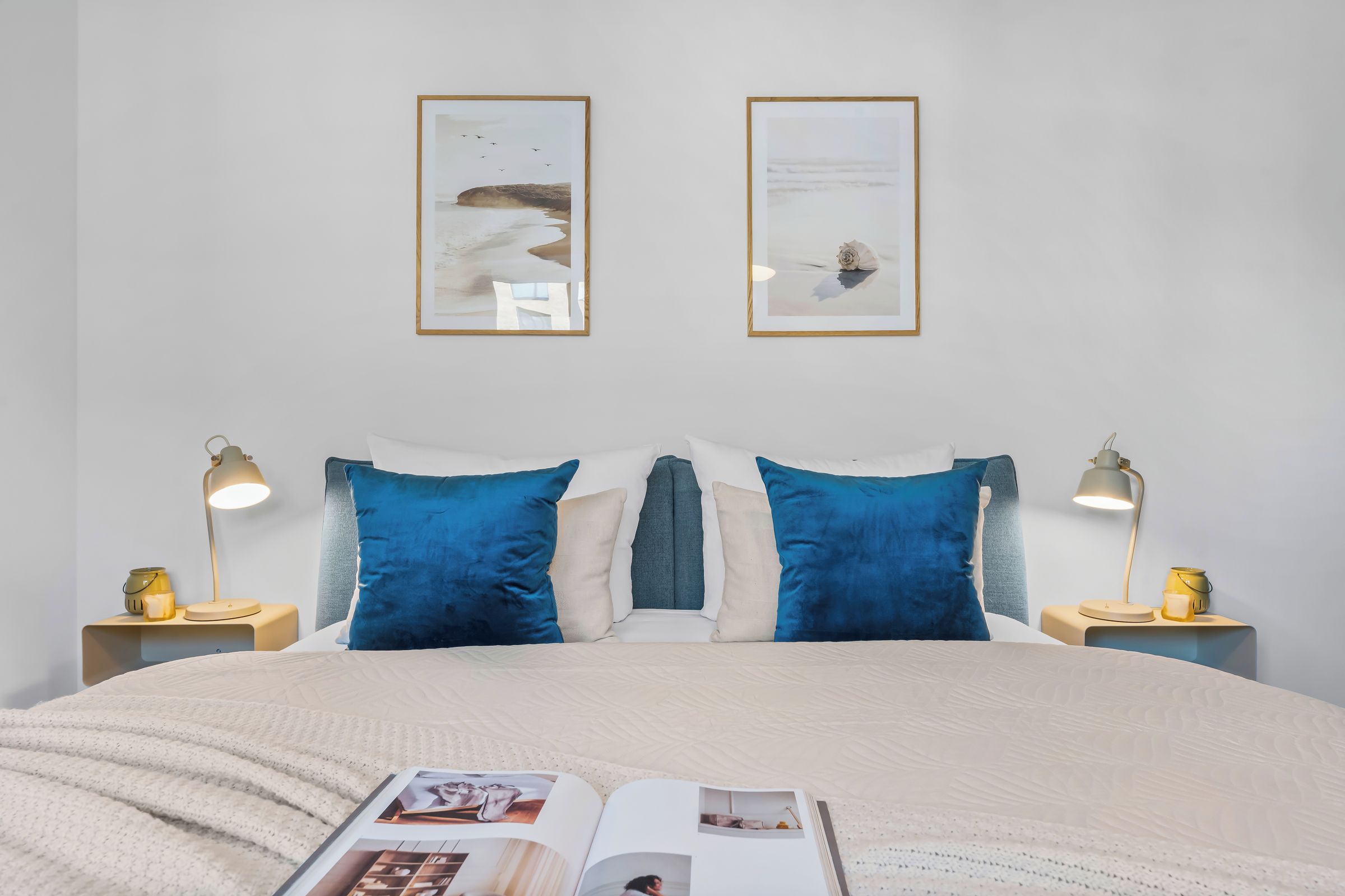 Un dormitorio con una cama, dos mesitas de noche, lámparas, un libro y dos cuadros en la pared.