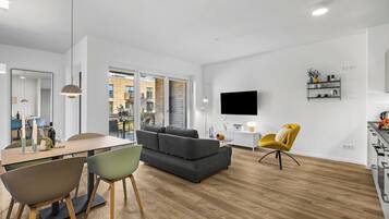 Ein modernes Wohnzimmer mit einem grauen Sofa, einer Essecke mit Tisch und Stühlen, einem gelben Sessel und einem an der Wand befestigten Fernseher.