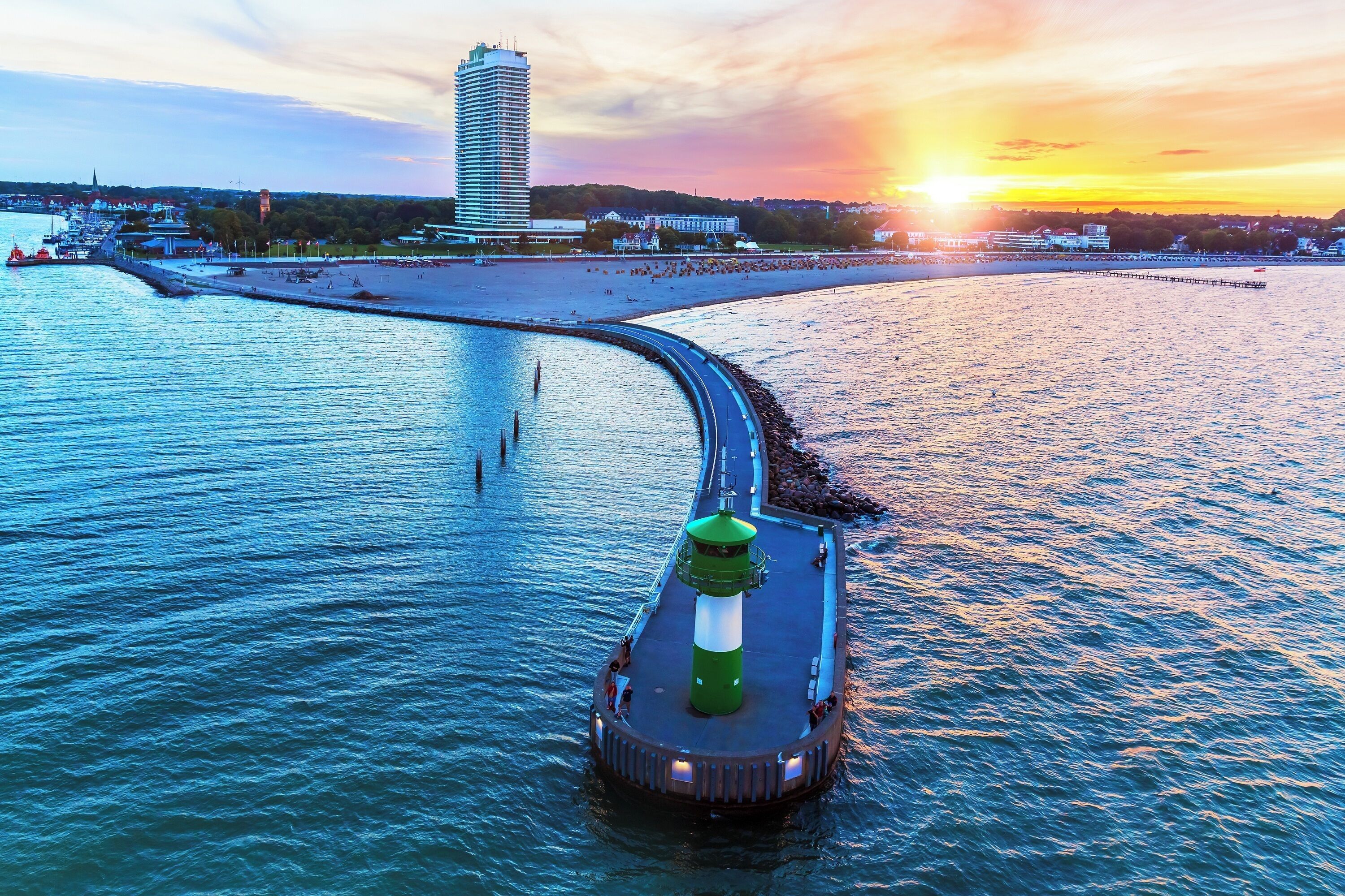 Escena costera con un faro en un muelle, un espigón que se adentra en el agua y un paisaje urbano al fondo.