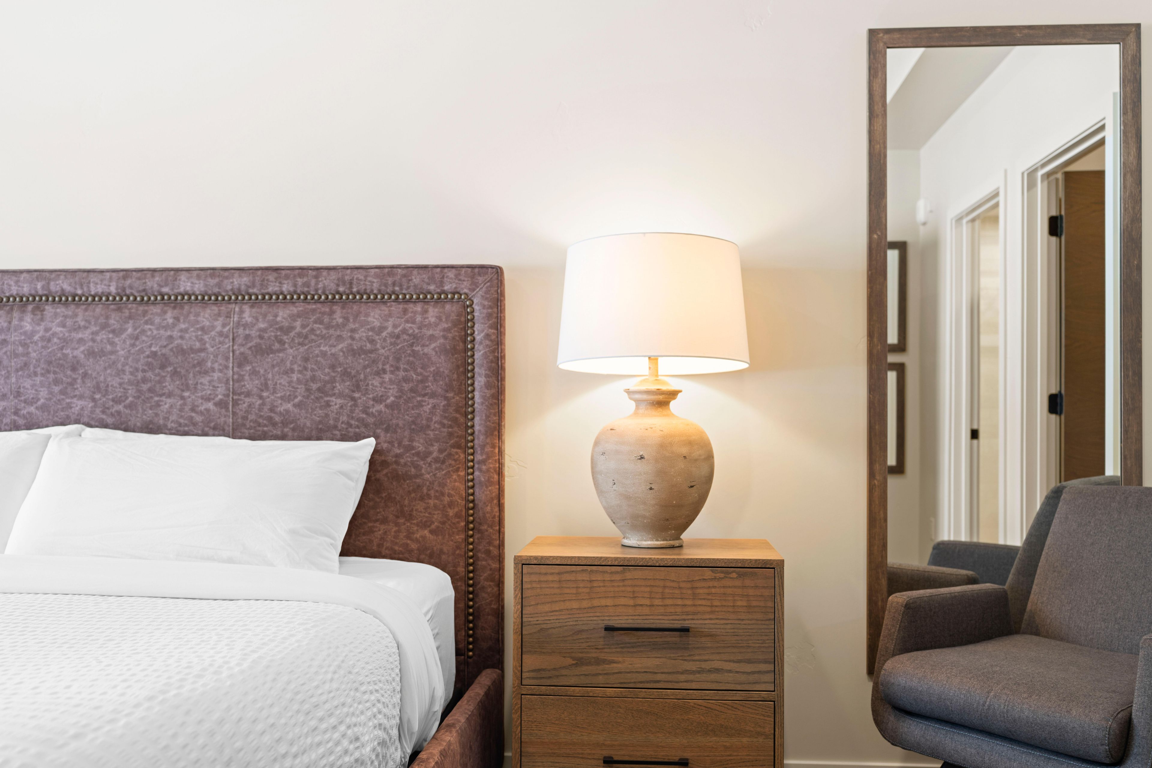 A neatly made bed with white linens, a wooden bedside table with a lamp, and a chair.