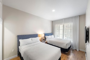 A hotel room with two beds, a bedside table with a lamp, a TV mounted on the wall, and a window with sheer curtains.