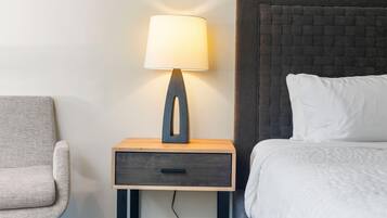 A hotel room with a bed, a nightstand with a lamp, a chair, and a wall with a textured headboard.