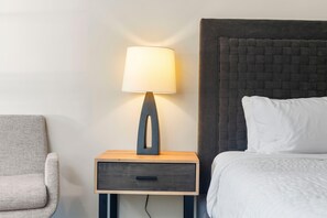 Habitación de hotel con cama, mesita de noche con lámpara, una silla y cabecero texturizado en la pared.