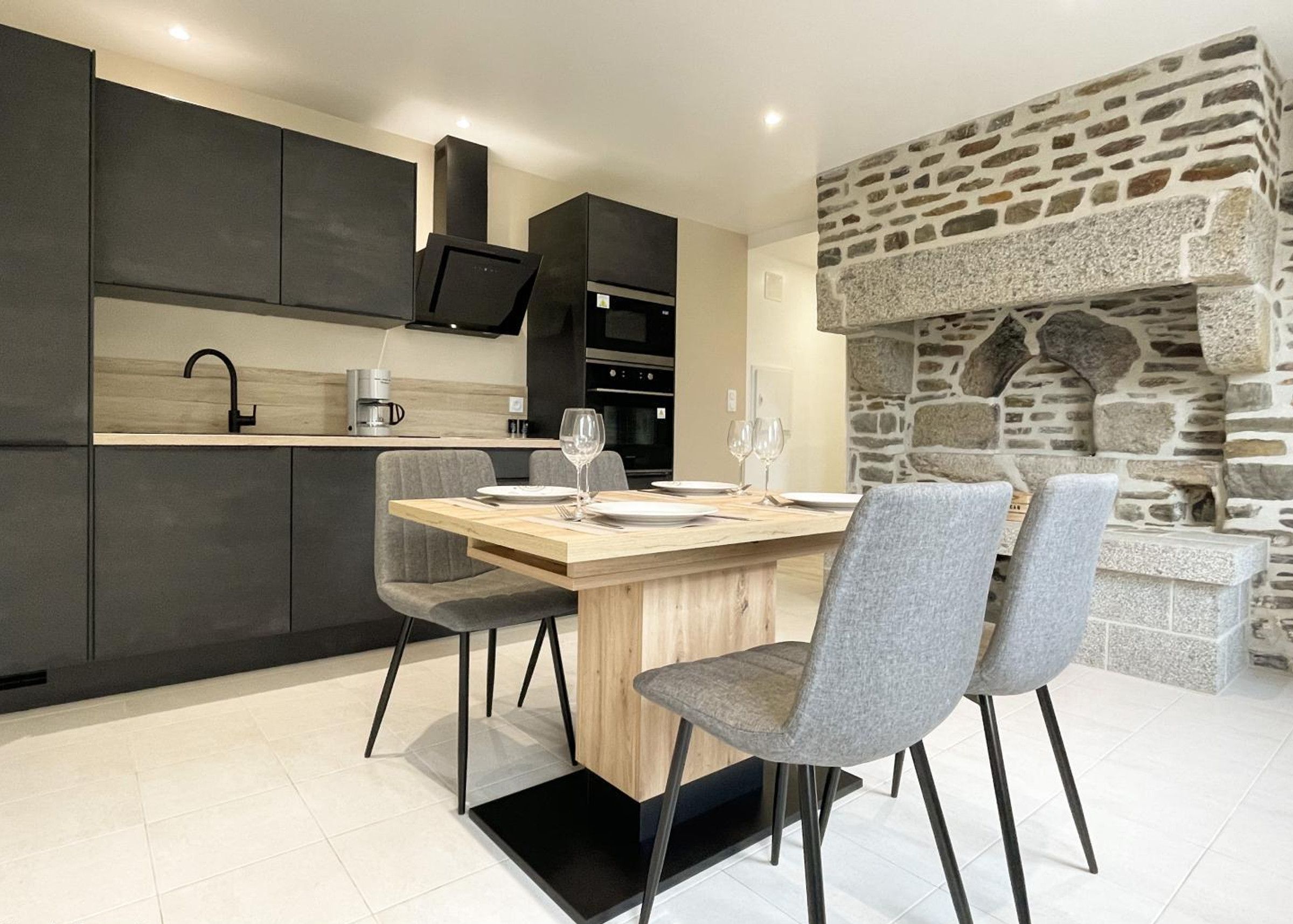 Una cocina moderna con armarios oscuros, una chimenea de piedra y un comedor con sillas grises y una mesa de madera.