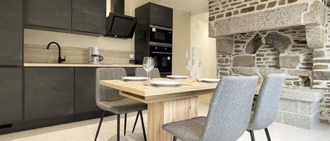 Une cuisine moderne avec des meubles de couleur sombre, une cheminée en pierre et un coin repas avec des chaises grises et une table en bois.