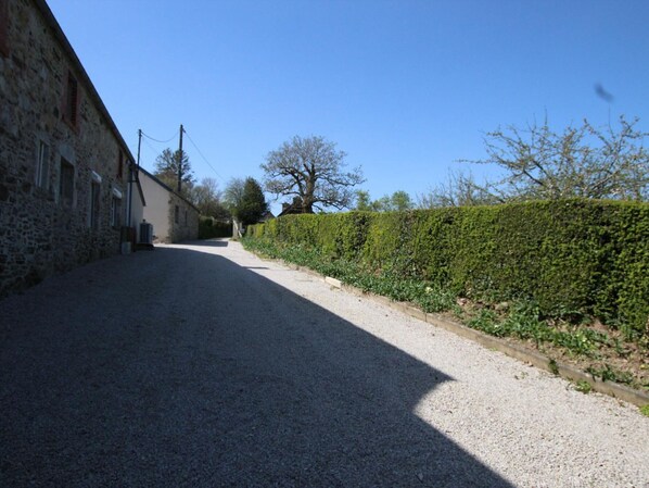 A gravel road leading to a stone building with a hedge on one side.