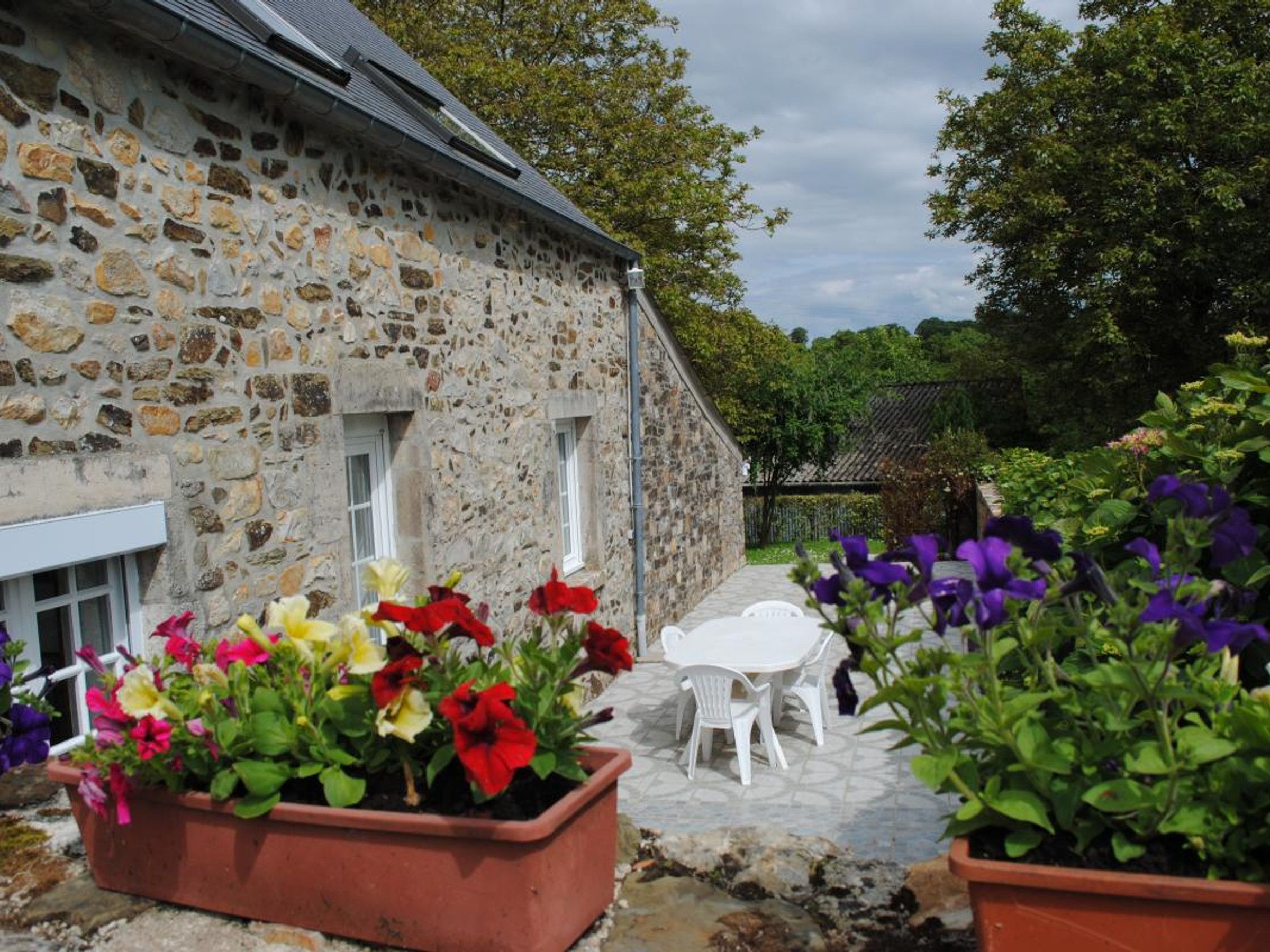 A stone building with a small patio area, white outdoor furniture, and potted plants with colorful flowers.