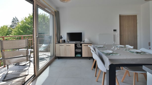 Ein moderner Essbereich mit einem großen Tisch, gedeckt für sechs Personen, einem Fernseher und einer Glastür, die auf einen Balkon führt.