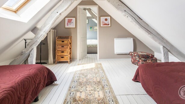 Une chambre confortable située sous les toits, avec un plafond en pente, des poutres en bois, un puits de lumière, une grande fenêtre, un lit, une commode et un radiateur.