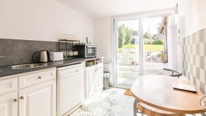 Une cuisine moderne équipée d’un lave-vaisselle, d’un four à micro-ondes et d’une bouilloire, une table ronde en bois et une porte-fenêtre coulissante donnant sur une terrasse avec coin salon extérieur.