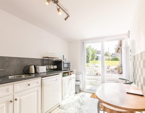 A modern kitchen with a dishwasher, microwave, and kettle, a round wooden table, and a sliding glass door leading to a patio with outdoor seating.