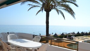 Ein Balkon mit weißem Tisch und Stühlen mit Blick auf einen Strand mit Sonnenschirmen und einer Palme.