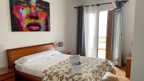 Ein Hotelzimmer mit Bett, Nachttisch, Lampe, einem Gemälde an der Wand und einem Balkon mit Vorhängen.