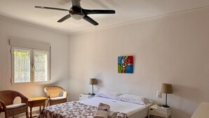 Habitación de hotel con cama, dos mesitas de noche, ventilador de techo, ventana con vistas, una silla y un cuadro en la pared.