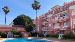 A swimming pool with a clear blue water surface, surrounded by a pink building with balconies and palm trees. - Penthouse in Denia with air conditioning and private parking (Denia)