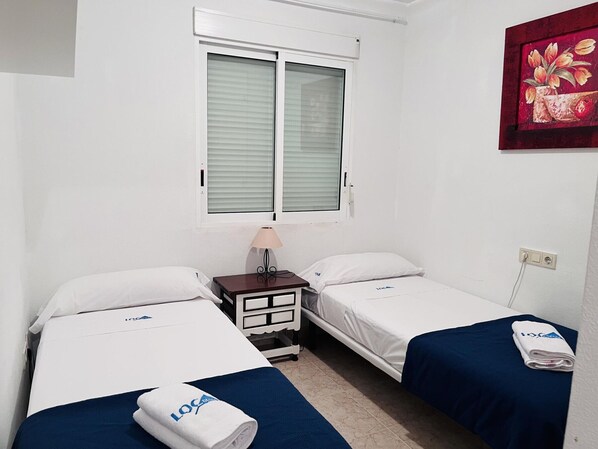 A hotel room with two beds, a nightstand, a lamp, a window with blinds, and a painting on the wall.