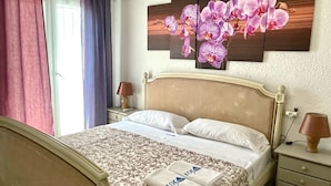 A hotel room with a bed, a nightstand, a wall-mounted orchid painting, and a window with curtains. - Cozy 2-bedroom bungalow in Els Poblets with Wi-Fi and parking (Denia)