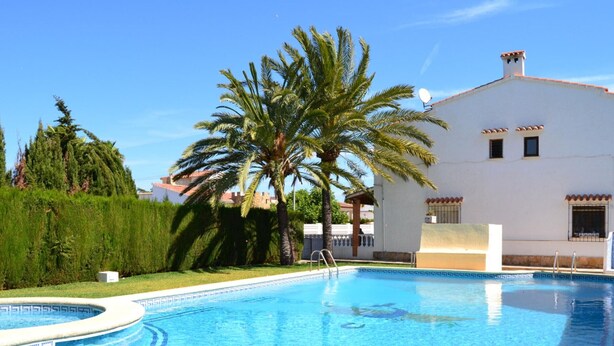 A clear blue swimming pool with a palm tree and a white building in the background. - Cozy 2-bedroom bungalow in Els Poblets with Wi-Fi and parking (Denia)