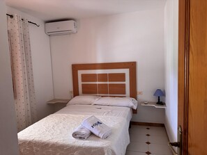 Ein kleines, sauberes Zimmer mit einem Einzelbett, einem Nachttisch, einer Lampe und einer Klimaanlage.
