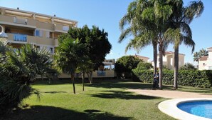 A pool area with a well-maintained lawn, palm trees, and a building with balconies in the background. - Charming 3-bedroom apartment with terrace and garden access in Denia (Denia)